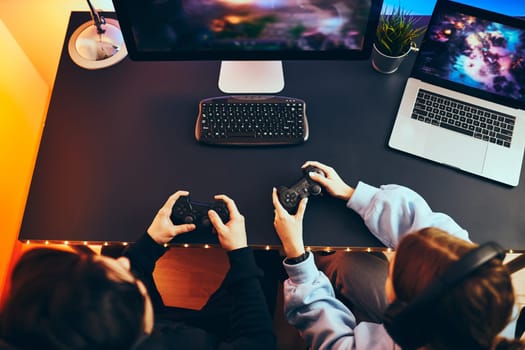 Friends playing video game at home. Gamers holding gamepads sitting at front of screen. Streamers girl and boy playing online in dark room lit by neon lights. Competition and having fun