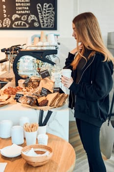 Woman holding cup with coffee looking at pastry, buns, cakes and cookies and waiting for the order. Girl buying a sweet food and hot drink to go. Young woman having a break doing shopping in a coffee shop