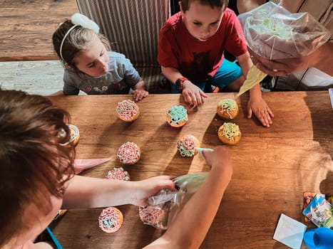 Group of children baking cupcakes, squeezing cream from confectionery bag, preparing ingredients, topping, sprinkles for decorating cookies. Kids cooking, working together in kitchen at home. Concept of happy family