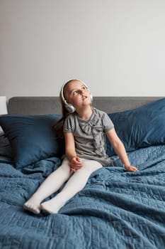 Little girl singing holding headphones cord imitating herself a real singer. Child having fun jumping dancing listening to music on bed in bedroom at home