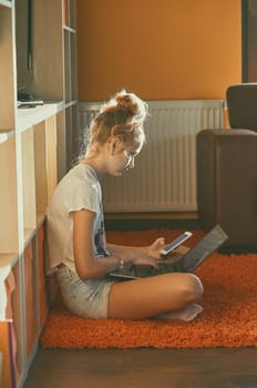Young woman using portable computer and mobile phone, sitting on a floor, learning online at home. Candid people, real moments, authentic situations