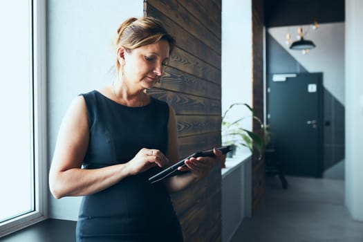 Businesswoman working on tablet in office. Mature woman using touch pad computer standing by window in modern interior. Female manager focused on work holding digital device. Using technology. Open office. Copy space right