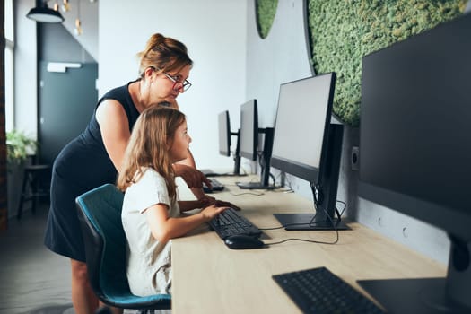 Computer class at school. Teacher assisting schoolgirl while class at primary school. Child learning computer on elementary computer science lesson. Teacher helping to find task solution. Giving clues. Smart little girl learning online at school. Back to school