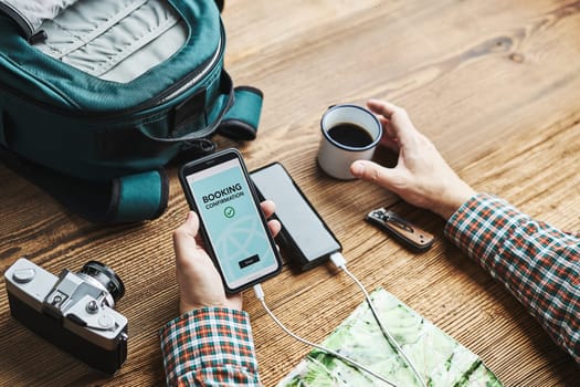 Man planning next vacation trip. Booking hotel online using smartphone. Ordering service. Charging mobile phone with power bank. Using technology while travelling. Man sitting at wooden table with camera and backpack drinking coffee. Travel plan concept