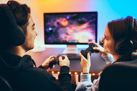 Friends playing video game at home. Gamers holding gamepads sitting at front of screen. Streamers girl and boy playing online in dark room lit by neon lights. Competition and having fun