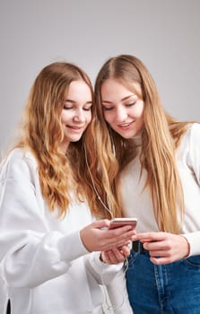 Young women girls listening to music together streaming content having fun watching video enjoying video chat talking with friends making gestures faces using smartphone earphones headphones standing over plain grey background