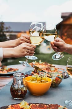 Friends making toast during summer picnic outdoor dinner in a home garden. Close up of people holding wine glasses with white wine over table with pizza, salads and fruits. Dinner in a orchard in a backyard