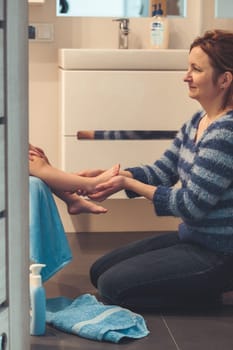 Mother applying moisturizing cream on her daughter's legs after bath. Mom caring about her child. Girl sitting in bathroom, wearing bathrobe
