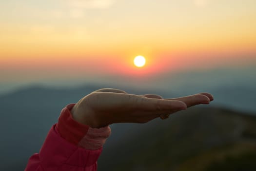 Sun over female hand. Sunrise in mountains. Ready for adventure. Natural mountain landscape with illuminated misty peaks, foggy slopes and valleys, blue sky with orange yellow sunlight
