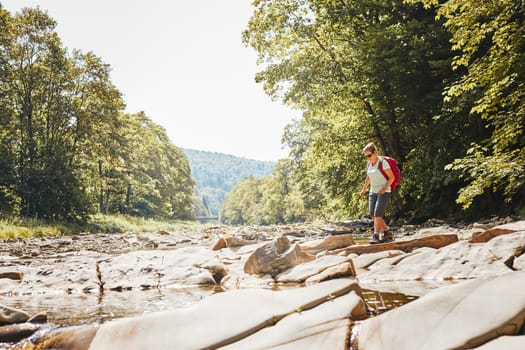 Trekking with backpack concept image. Female backpacker wearing trekking boots crossing mountain river. Woman hiking in mountains during summer trip. Vacation trip close to nature. Natural scenery