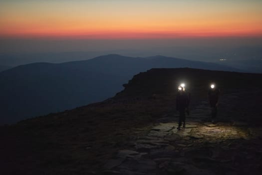 People hiking at night to see sunrise. Mountains at sunrise. Man standing on peak. Natural mountain landscape with illuminated misty peaks, foggy slopes and valleys, blue sky with orange yellow sunlight. Amazing scene from Beskid Zywiecki in Poland