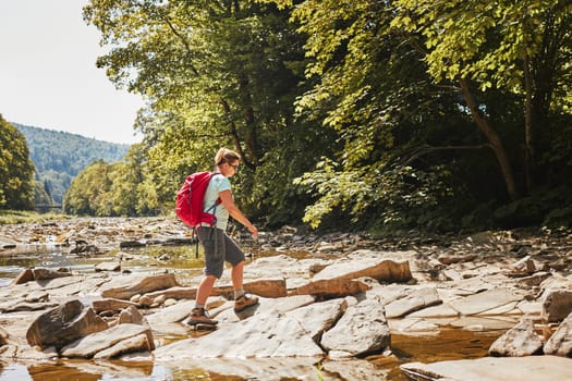Trekking with backpack concept image. Female backpacker wearing trekking boots crossing mountain river. Woman hiking in mountains during summer trip. Vacation trip close to nature. Natural scenery