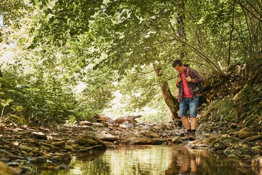 Trekking with backpack concept image. Backpacker in trekking boots crossing mountain river. Man hiking in mountains during summer trip. Vacation trip close to nature. Natural scenery