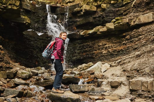 Hiking in mountains. Woman enjoying hike on sunny vacation day. Female with backpack walking close to waterfall. Spending summer vacation close to nature