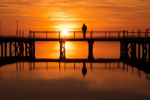 A person stands alone on a pier at sunset, reflecting on the beauty of the seascape before them. The warm orange hues of the sky create a sense of calmness and reflection, with the figure cast in silhouette against the beauty of the scenery. This scene can evoke feelings of introspection and appreciation for the natural world. (generative AI)