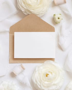 Blank card and envelope near cream roses and white silk ribbons top view, mockup. Romantic wedding scene with horizontal card and pastel flowers flat lay. Valentines or Spring concept