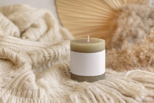 Candle with label on cosy sweater near dry pampas grass and palm leaf Close up, copy space. Boho packaging mock up. Home decorations, Bohemian, ethnic or scandinavian style