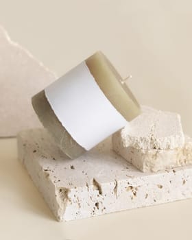 Candle with label on beige near cream travertine stones, Close up, copy space. Brand packaging mock up. Home decoration, minimal interior, Bohemian or scandinavian style