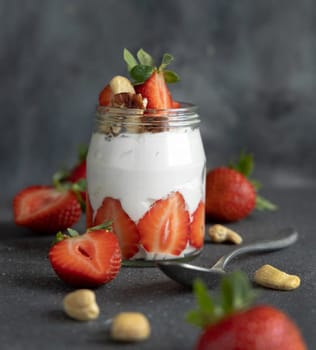 Greek yogurt, nuts and strawberries in a glass jar on grey concrete table with a spoon, close up. Healthy breakfast with fruits and nuts. Great way to eat protein