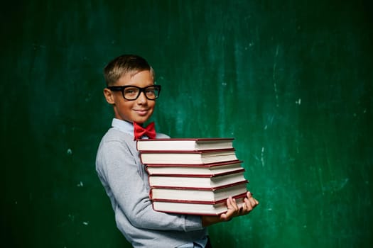 Cute schoolboy holding stack of books against blackboard