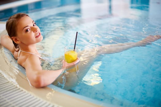 Young female in bikini holding glass of juice while relaxing in swimming pool