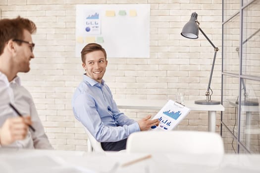 Happy businessman with financial document pointing at it while looking at his co-worker