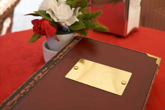 Detail of leather bound menu with blank name plate on red table cloth. There's a small flower vase on the side with red and white roses.