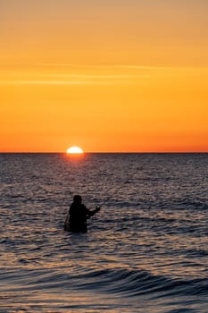The silhouette of an angler in waterproof trousers fishing in front of a dreamy orange sunset in the Baltic Sea. He stands in the middle of the water of the Baltic Sea.
