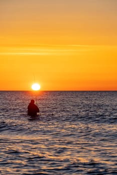 The silhouette of an angler in waterproof trousers fishing in front of a dreamy orange sunset in the Baltic Sea. He stands in the middle of the water of the Baltic Sea.