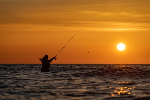 The silhouette of an angler in waterproof trousers fishing in front of a dreamy orange sunset in the Baltic Sea. He stands in the middle of the water of the Baltic Sea.