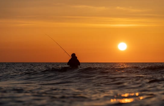 The silhouette of an angler in waterproof trousers fishing in front of a dreamy orange sunset in the Baltic Sea. He stands in the middle of the water of the Baltic Sea.