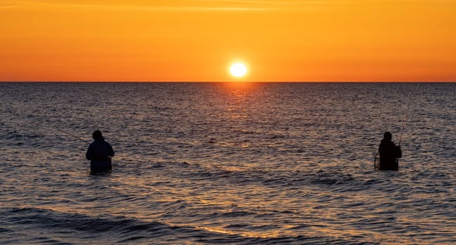 The silhouettes of two anglers in waterproof trousers fishing in front of a dreamy orange sunset in the Baltic Sea. They are standing in the middle of the water of the Baltic Sea.