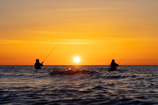 The silhouettes of two anglers in waterproof trousers fishing in front of a dreamy orange sunset in the Baltic Sea. They are standing in the middle of the water of the Baltic Sea.