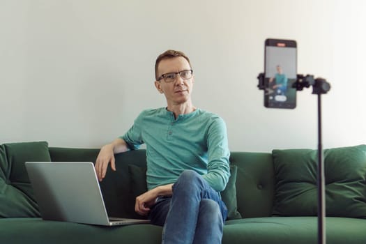 Handsome man, blogger, psychologist, sits at home on couch in front of phone camera and blogs, shoots videos, records interviews. Modern technologies. Online training. Video recording of trainings.