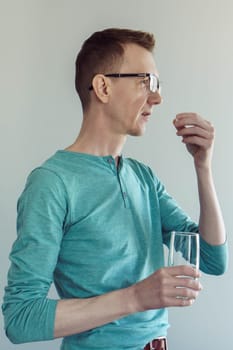 Close-up portrait of man in turquoise cotton T-shirt and glasses, standing on light background with glass of water and drinking pill, vitamins, antidepressants. Painkillers. Gastroenterology.