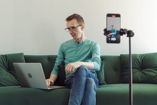Man in a T-shirt and jeans is sitting at home on couch in front of phone camera and typing something in notebook. Modern technologies. Online training. Male blogger. Video chat.