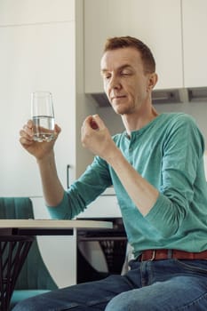 Lonely man is sitting at table at home against background of kitchen holding glass of water in his hand and is about to take pill. Feeling unwell. Painkillers. Vitamins. Antidepressants.
