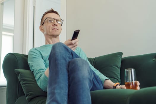 Handsome man in comfortable home clothes is sitting at home on green sofa with TV remote control in his hands and is looking for interesting channel. Free time and rest. Home life. Lonely people.