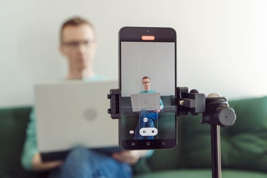 Focus on image from camera on phone screen, male blogger is sitting on couch at home against light wall and filming his interview. Modern technologies. Online training. Video recording of trainings.