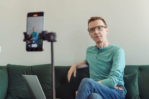 Male blogger. Video chat. Handsome man, psychologist, teacher sits at home on couch with laptop in front of phone camera and shoots video. Modern technologies. Video recording of trainings.