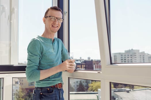 Positive emotions. Man in turquoise T-shirt with long sleeves stands on balcony of house, apartment, office with glass of juice and smiles, looking at camera. City and buildings. Tourism and travel.