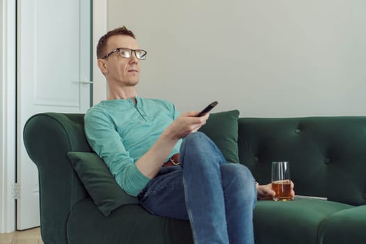 Man with glasses for vision in comfortable home clothes is sitting at home on green sofa with TV remote control in his hands, glass of beer is looking for interesting channel. Free time and rest.