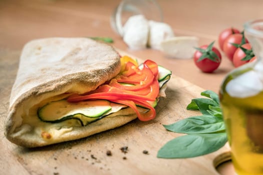 Italian food, closed calzone pizza on rustic wooden background. Filled with peppers and zucchini. On blurred background small red tomatoe , mozzarella and crude oil. In the foregroud we also see basil. Selective focus