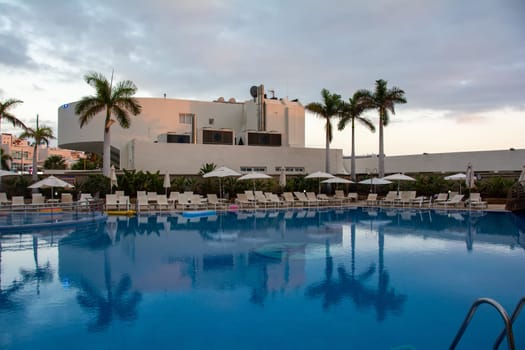 A swimming pool with palm trees, hotel and sun loungers in the late afternoon