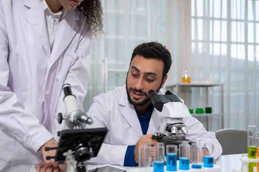 Male scientist in white laboratory coat discussing with young female assistant for data analysis on tablet while working with microscope for research in scientific research laboratory