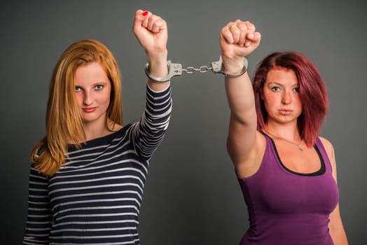 Two angry women connected by a pair of handcuffs, fist, blond and red-haired, on grey background