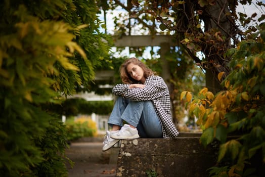 Beautiful young woman sitting on white wooden bench in tranquil garden with lush greenery, wearing casual attire including black and white checkered jacket, blue jeans, and white sneakers, showcasing peaceful natural atmosphere. The image captures a beautiful young woman sitting on a white wooden bench in a serene garden or park setting. She is wearing a black and white checkered jacket over a white shirt, paired with blue jeans and white sneakers. The background is lush with greenery, including various bushes and plants, creating a peaceful and natural atmosphere. Her face is intentionally blurred to maintain privacy, adding a sense of anonymity to the scene. The overall composition of the image exudes a casual and relaxed vibe, perfect for conveying tranquility and connection with nature. This image is ideal for themes related to outdoor living, mindfulness, casual fashion, and lifestyle branding. The natural elements and serene setting make it suitable for use in advertising, social media, blogs, and editorial content focused on relaxation, nature, and modern aesthetics.