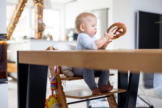 Happy infant sitting at dining table and playing with his toy in traditional scandinavian designer wooden high chair in modern bright atic home. Cute baby playing with toys.