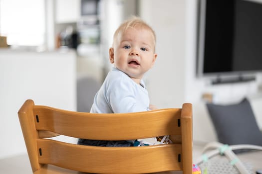 Happy infant sitting in traditional scandinavian designer wooden high chair and laughing out loud in modern bright home. Cute baby smile