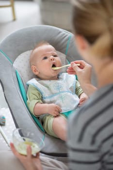Mother spoon feeding her baby boy child in baby chair with fruit puree. Baby solid food introduction concept